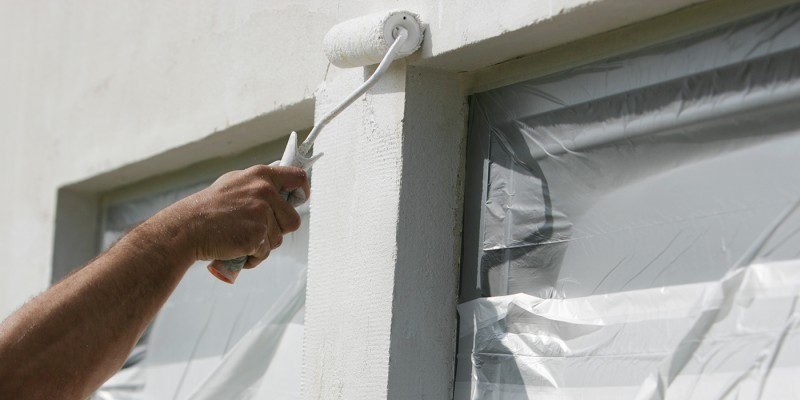 Handwerker streicht mit Farbrolle eine verputzte Fassade weiß, Fenster sind abgeklebt