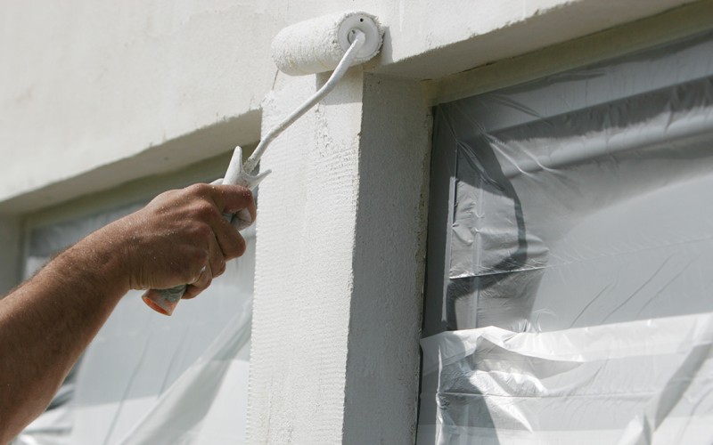 Handwerker streicht mit Farbrolle eine verputzte Fassade weiß, Fenster sind abgeklebt